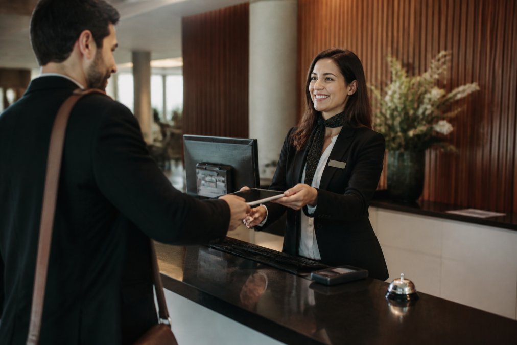 A hotel front desk manager in a modern lobby handing a tablet to a guest checking in, with a computer screen showing a booking management system in the background
