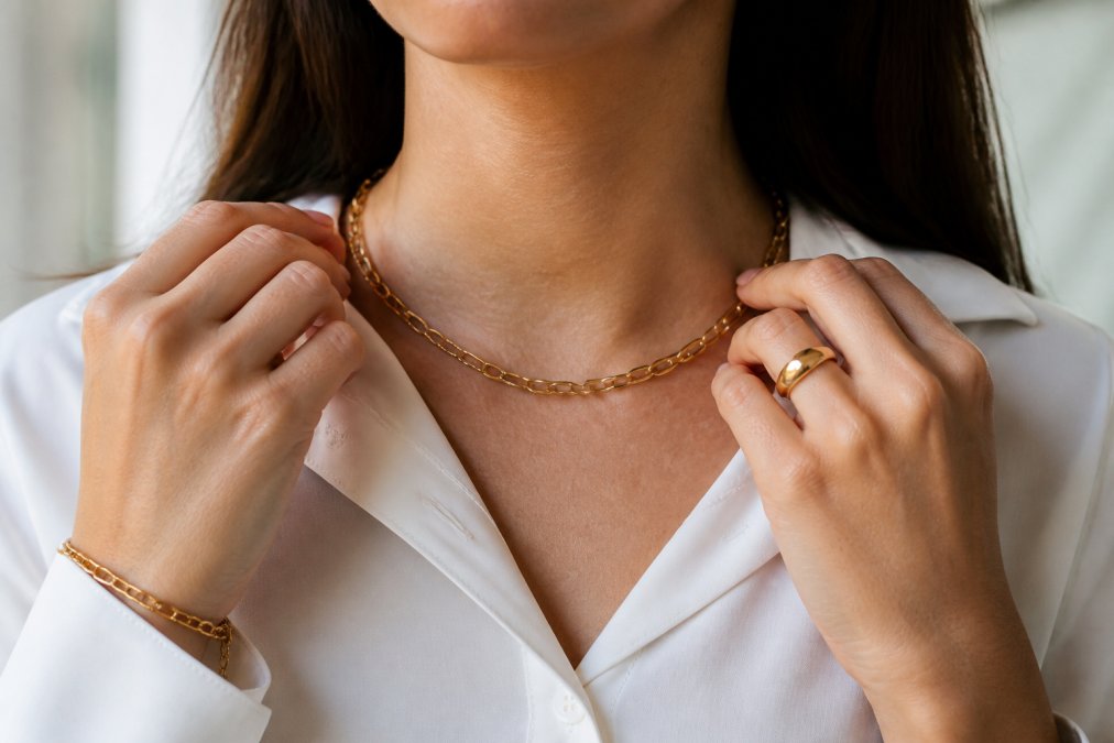 Close-up of hands adjusting a gold-plated chain necklace worn with a crisp white professional blouse, showing how jewelry adds polish to a minimal everyday outfit with natural lighting