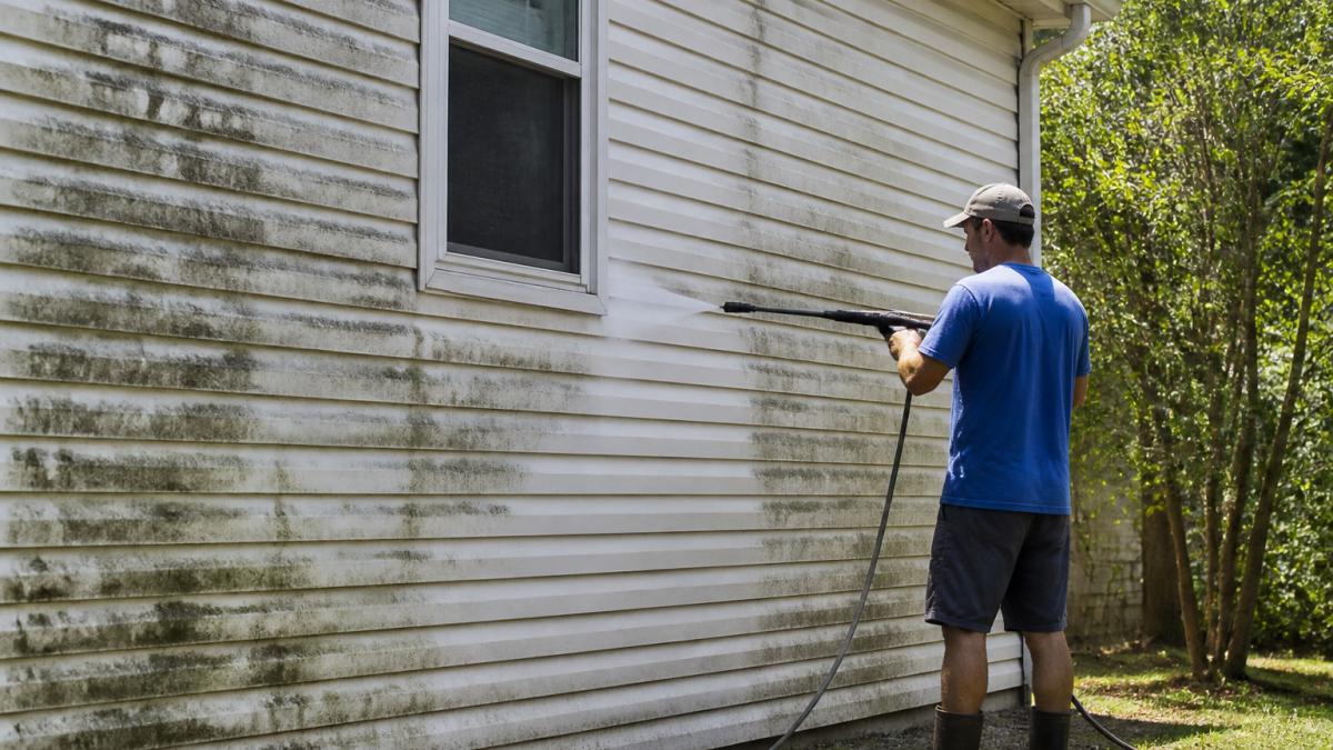 Professional contractor performing house washing on residential vinyl siding, demonstrating proper technique with visible dirt and algae being cleaned away, natural daylight illuminating the work area