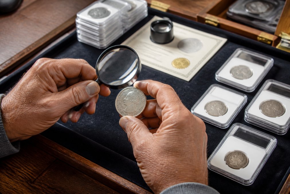 A close-up of a coin dealer's hands holding a magnifying glass over a rare coin on a felt tray, with a grading certificate and slabbed coins in the background