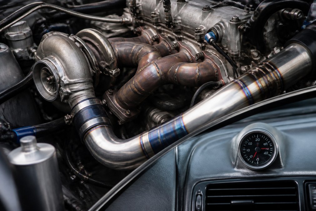 Close-up of an engine bay with turbocharger mounted on exhaust manifold, intercooler piping visible with heat-colored aluminum components, boost gauge showing pressure readout on dashboard