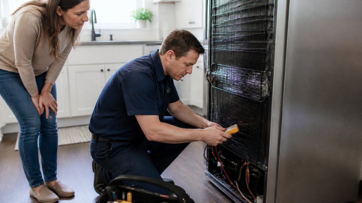 Professional appliance repair technician in uniform using diagnostic tools on a kitchen refrigerator while a homeowner observes in a bright, modern kitchen