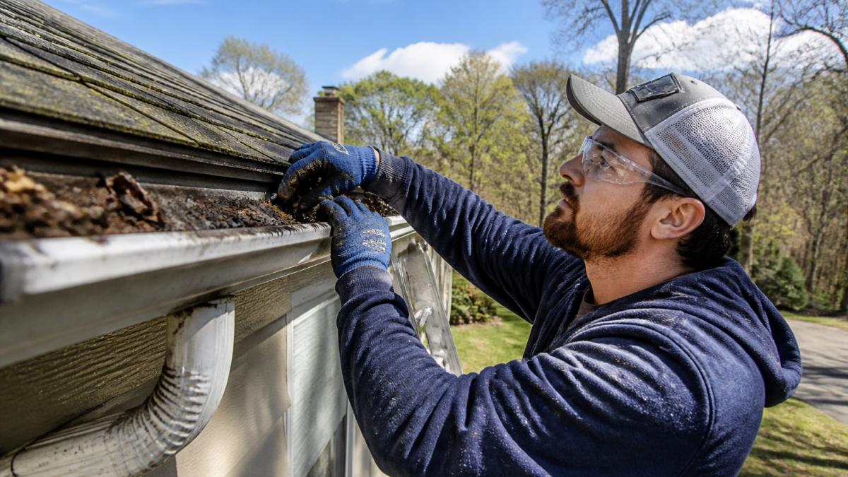 Homeowner in safety gear and work gloves inspecting roof gutters and downspouts during spring maintenance, with clear sky and house exterior visible