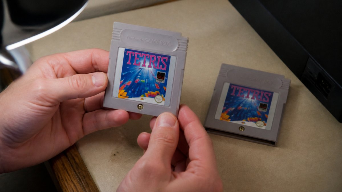 Close-up of a gamer's hands holding a Game Boy cartridge under a desk lamp, comparing the label and screw to an authentic copy on the table beside it