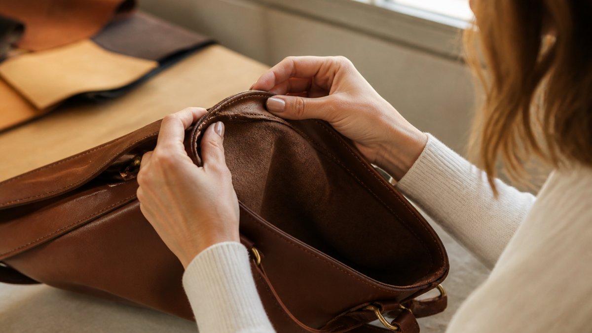 Close-up of a woman's hands turning a leather handbag inside out to examine the stitching along the seam, soft natural window light, neutral studio background