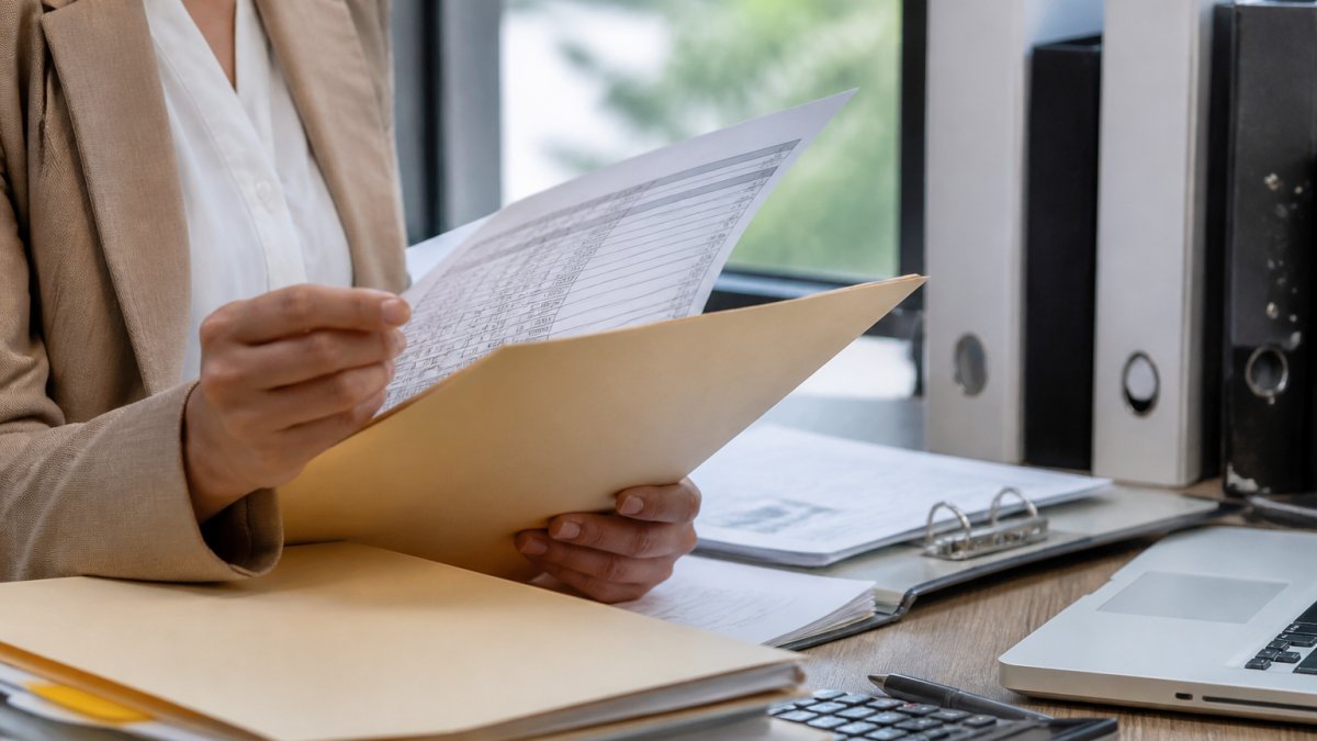 Professional HR manager organizing workers' compensation payroll records at desk with folders, documents, and computer displaying payroll data in organized workspace