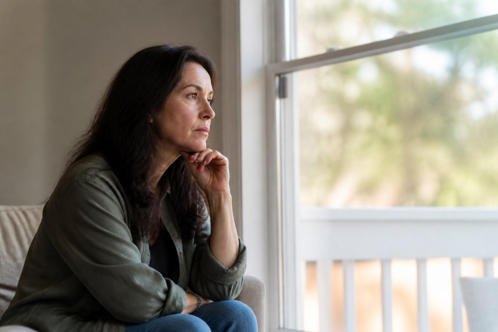 Adult sitting thoughtfully by a large window with natural light, hand to chin, gazing outward with a contemplative expression suggesting emotional weight and internal struggle