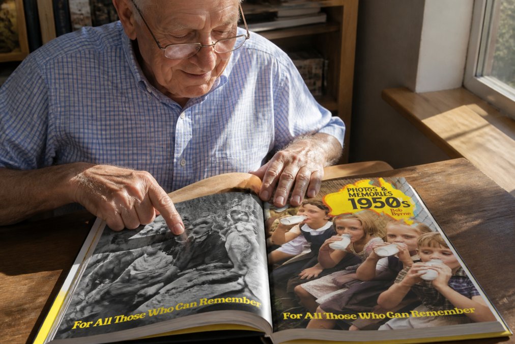 An older adult with reading glasses carefully examining an open history photo book at a wooden table, pointing to a photograph with a warm, engaged expression, natural afternoon light streaming across the pages