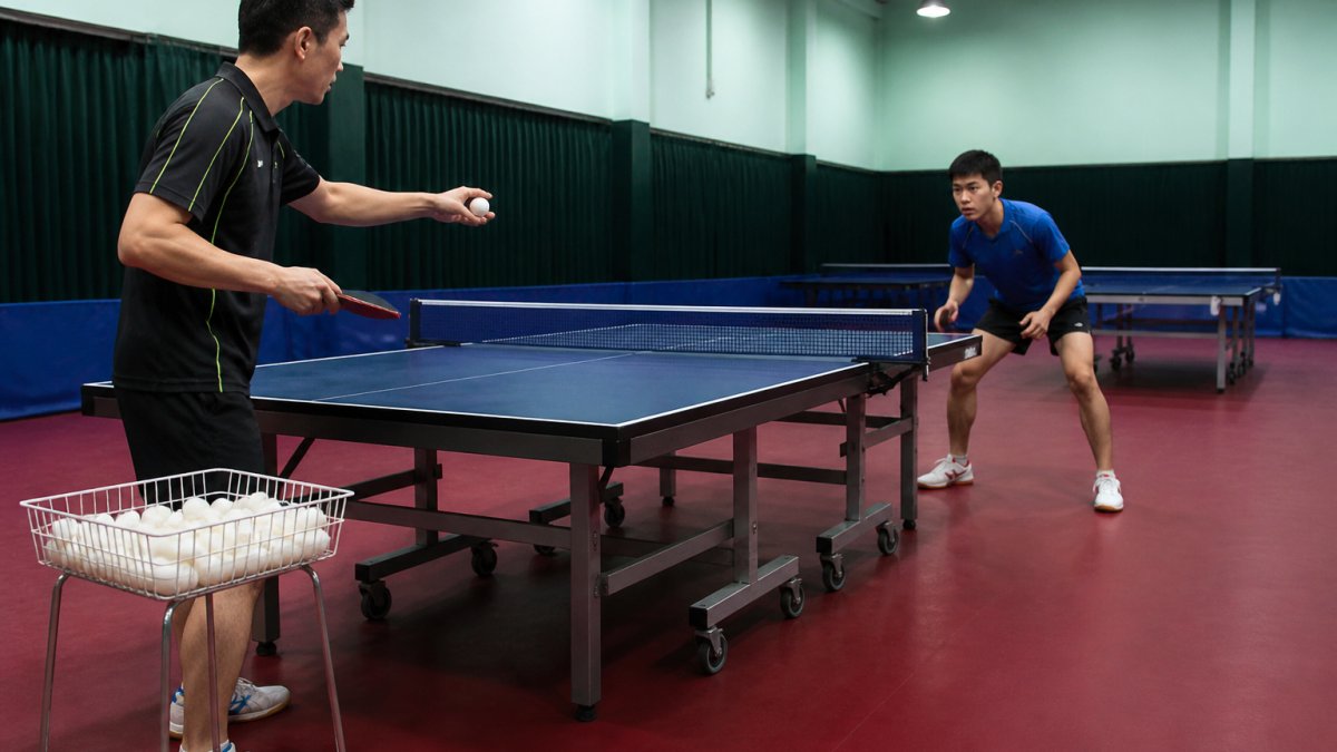 A table tennis coach standing beside a table holding a racket and a ball in feeding position, with a basket of balls nearby and a player ready in a ready stance across the table