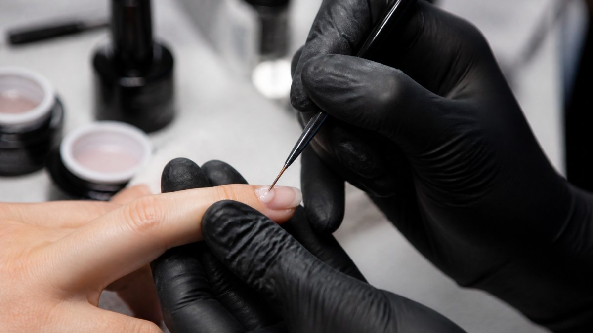 Nail technician using a fine brush to apply a bead of builder gel near the cuticle of a natural nail.