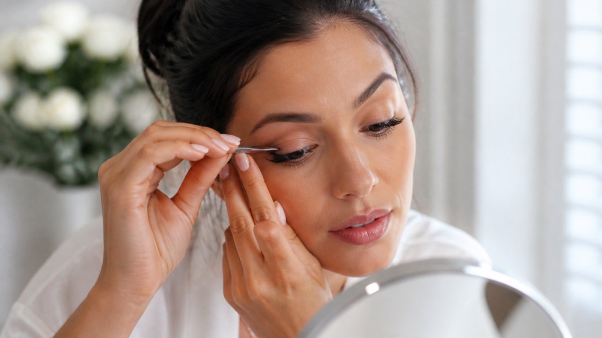 Close-up of a woman's hands using tweezers to press a false lash strip onto her lash line, looking down into a mirror in a bright vanity setting