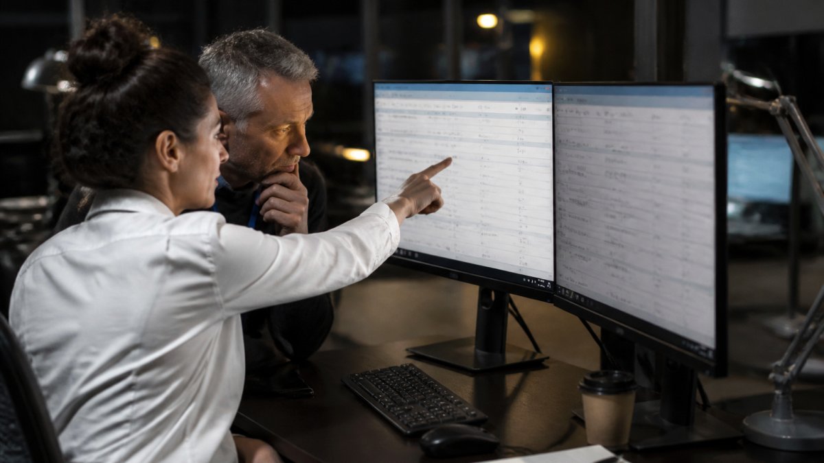 Two risk analysts at a large enterprise reviewing flagged AI agent decisions on dual monitors in a dim operations center, one pointing at a queue of approval items on screen
