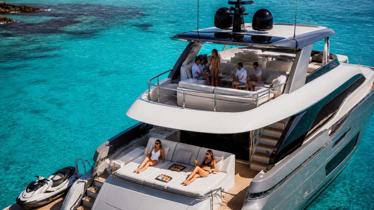 Aerial view of a luxury motor yacht anchored in clear turquoise waters with guests enjoying the flybridge and sun deck, with white loungers and cocktails visible