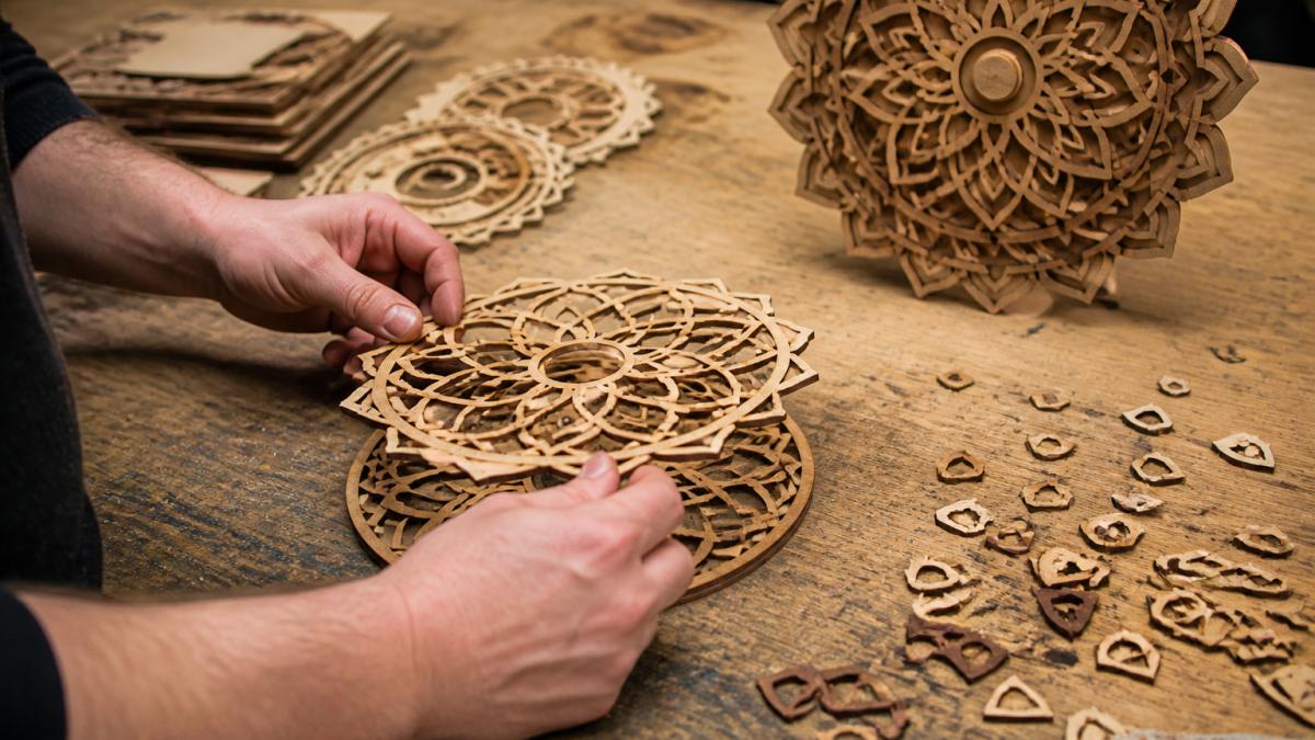 Close-up of a maker's hands stacking thin laser-cut wood mandala rings on a workbench, with a finished three-layer mandala wall piece and small wood scraps nearby