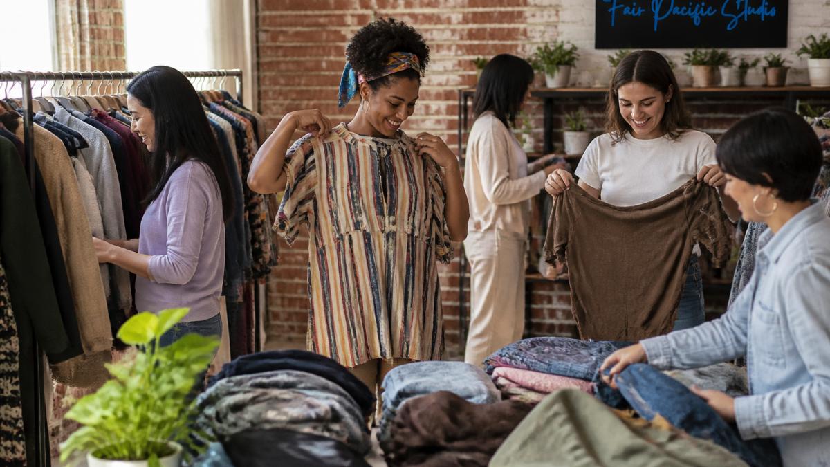 Group of diverse women browsing clothing items on racks and tables at a community clothing swap event, smiling and trying on pieces