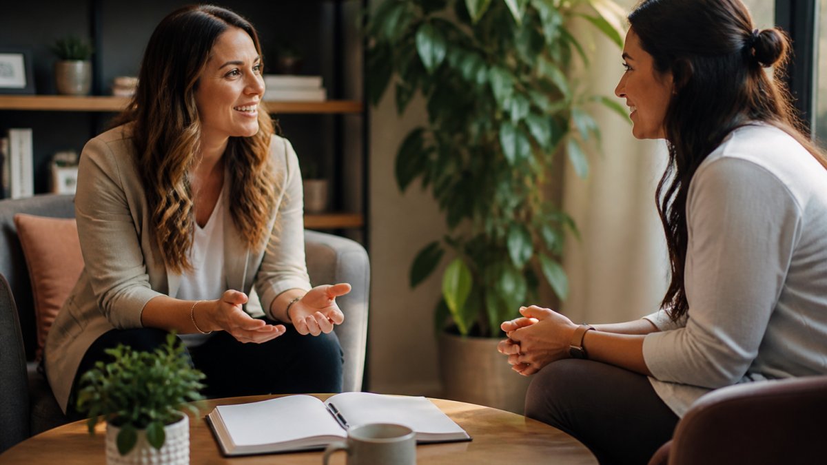 A coach and client sitting across from each other in a bright, comfortable office, engaged in a focused conversation with a notebook open on the table