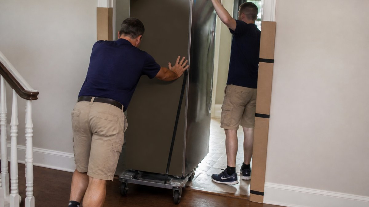 Two people use an appliance dolly to move a refrigerator through a narrow doorway protected by cardboard.