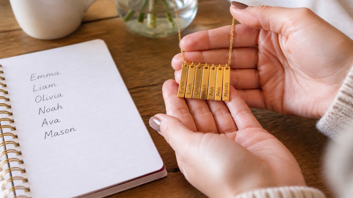 Close-up of a woman's hands holding a gold name necklace with several engraved names beside a handwritten list of family names on a notepad, warm natural light on a wooden table
