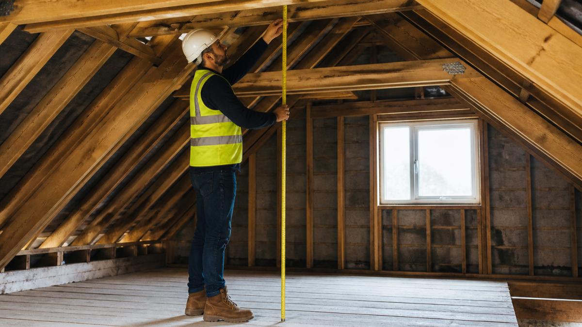A surveyor in a hard hat and high-visibility vest measuring head height with a tape in a bare, timber-framed loft space, roof trusses visible overhead and daylight coming through a small window