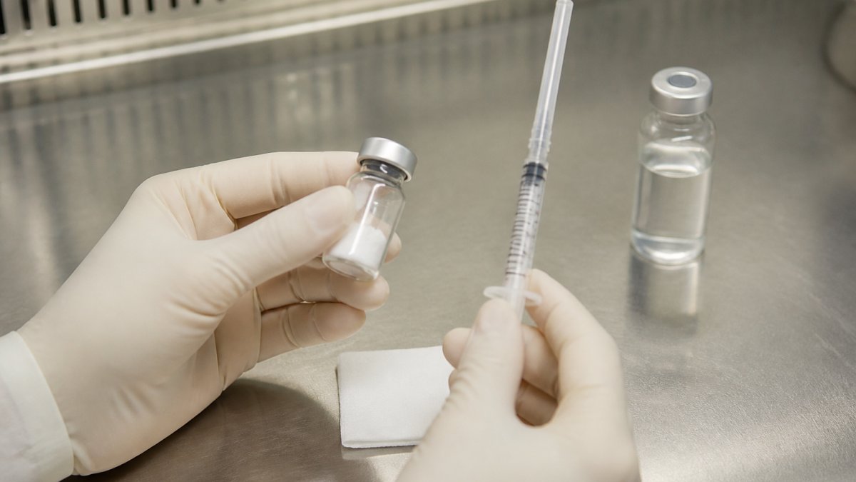 Close-up of gloved hands holding a small glass peptide vial and a mixing syringe beside an alcohol wipe on a clean stainless steel lab bench, with a second vial of clear liquid in the background