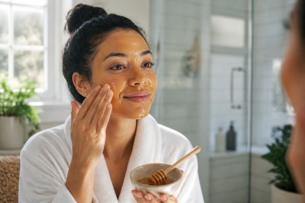 A woman in a bright bathroom applying a golden honey face mask with her fingertips, wearing a soft robe, warm natural light