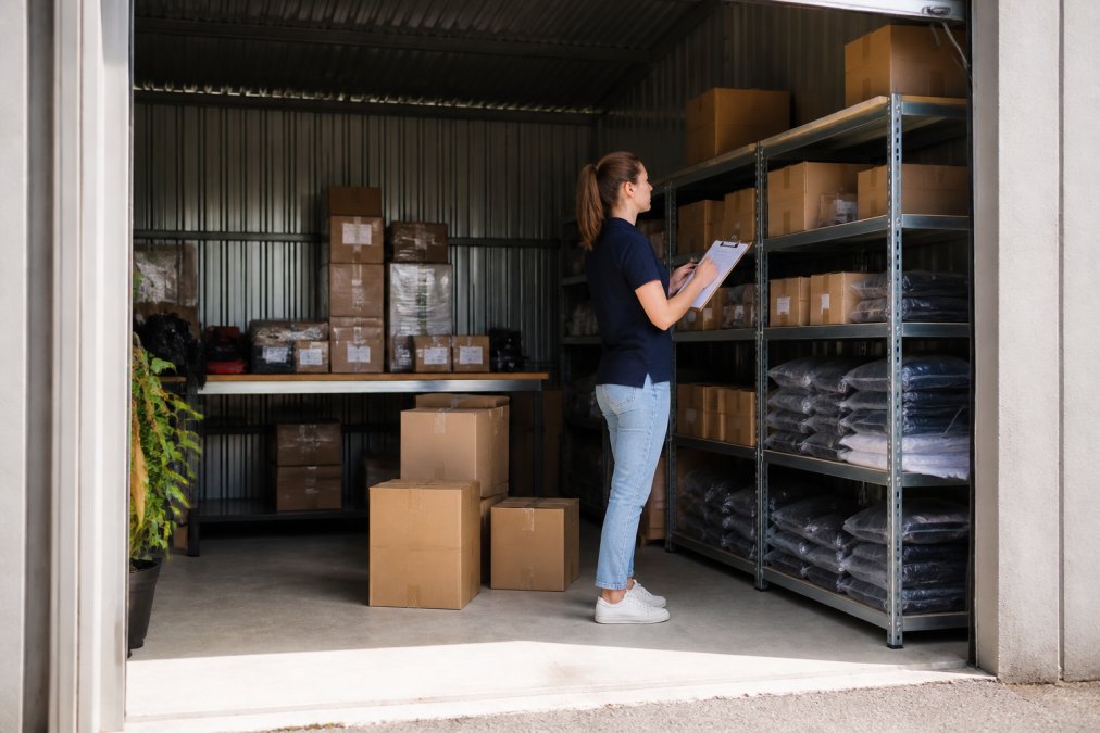 E-commerce business owner or team member inside a commercial storage unit, reviewing inventory on a clipboard, checking product labels on organised shelves, natural light from the open door illuminating the space
