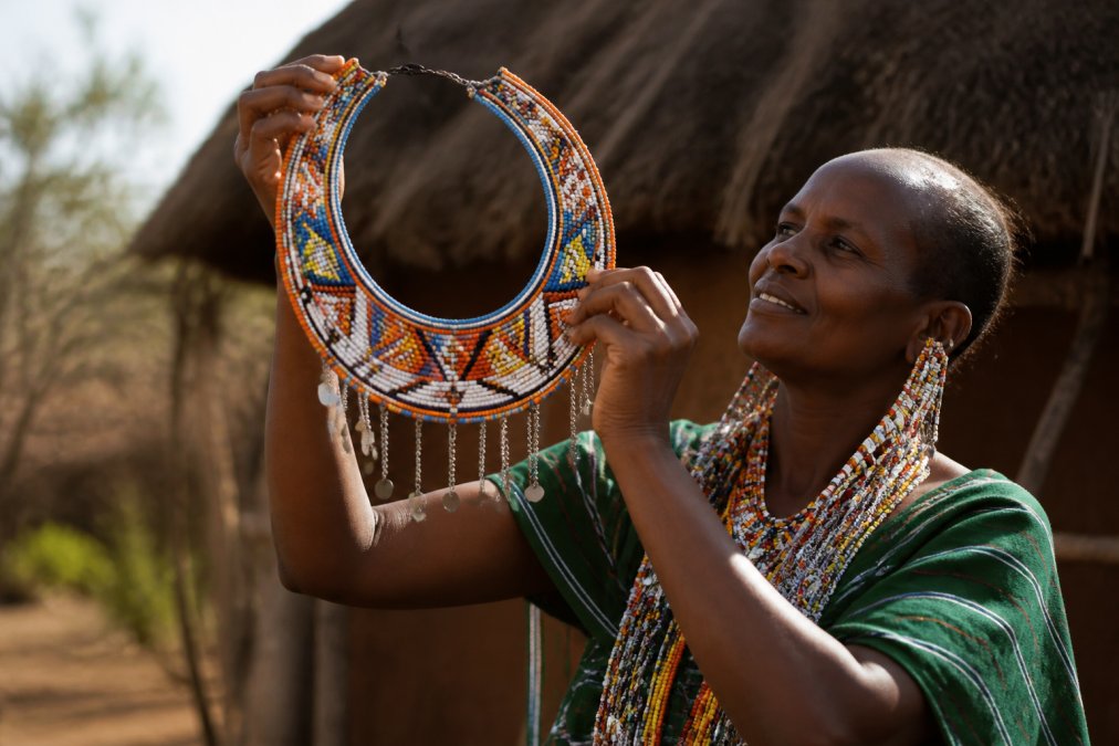 Skilled artisan displaying a finished handcrafted beaded necklace with traditional East African geometric patterns, pride evident in their expression, holding the piece up to natural light to show the intricate detail