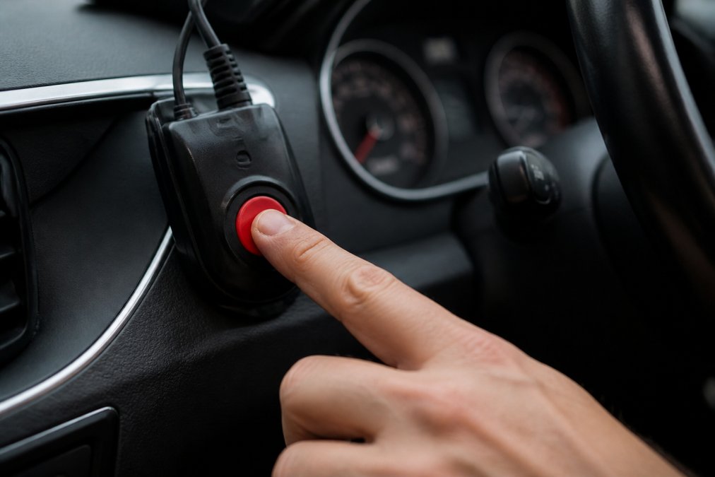How GPS Emergency Tracking Works: A 2026 Guide Close-up of a person's hand pressing a red SOS button on a compact GPS tracking device mounted inside a vehicle, the dashboard and motorway visible through the windscreen in soft natural daylight