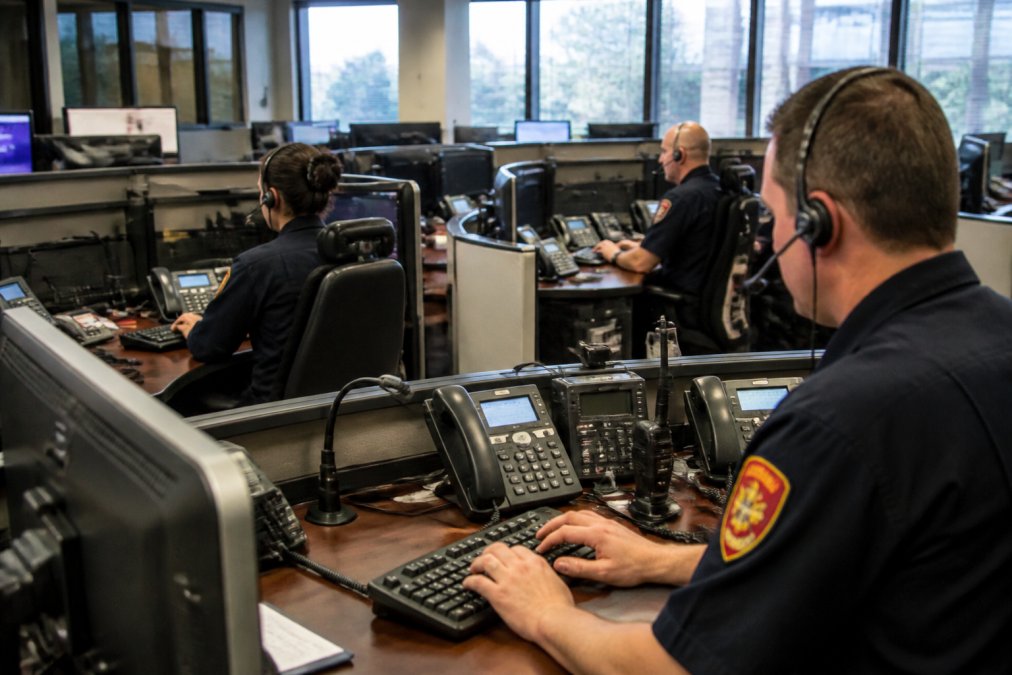How GPS Tracking Helps Emergency Services Emergency dispatch control room with multiple operators monitoring real-time vehicle locations displayed on large screens, with communication headsets and data terminals visible under bright fluorescent lighting