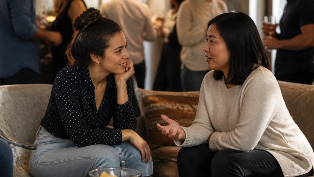 Two people having a genuine conversation at a social gathering, one person leaning in attentively while the other speaks, both appearing relaxed and engaged in a comfortable setting