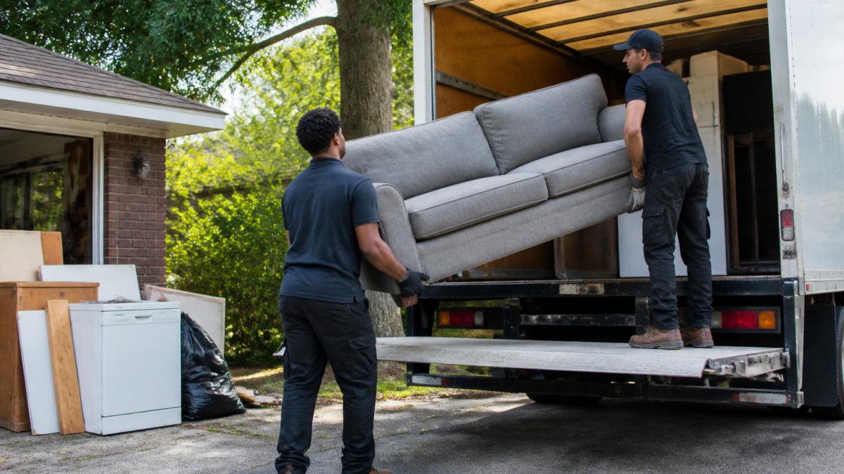 Professional junk removal crew loading furniture and appliances into the back of a truck at a residential property, showing two workers coordinating a large sofa being lifted into the truck bed