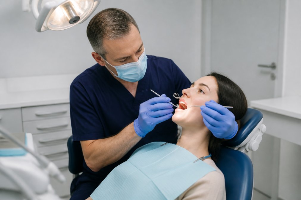 A dentist in blue gloves and a surgical mask examining a patient's teeth with a small mirror and probe, patient reclined in a modern dental chair under a bright overhead light in a clean, white-walled surgery