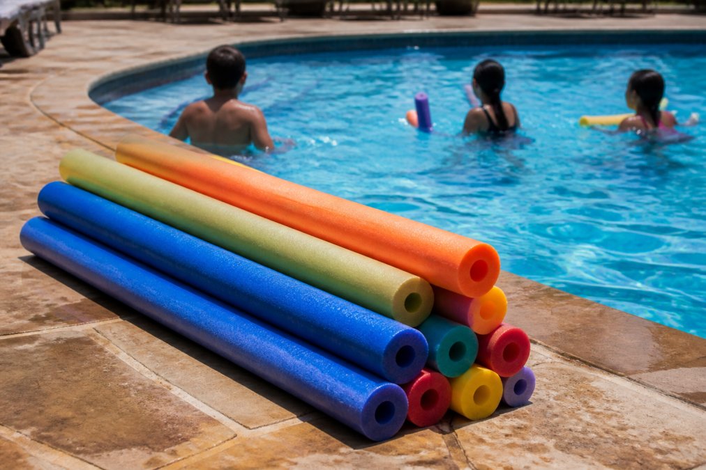 Colorful jumbo pool noodles of varying lengths stacked together poolside, showing size comparison with swimmers in background