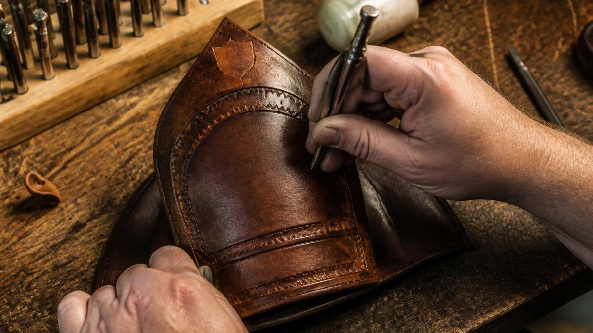 Close-up of a leather helmet front being hand-tooled on a wooden workbench, showing detailed craftsmanship with tooling marks and leather shaping