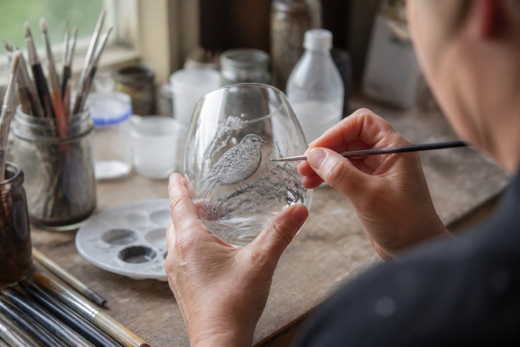 Artist's hands working on hand-painted glassware with a brush, showing detailed wildlife design in progress on a tumbler or wine glass