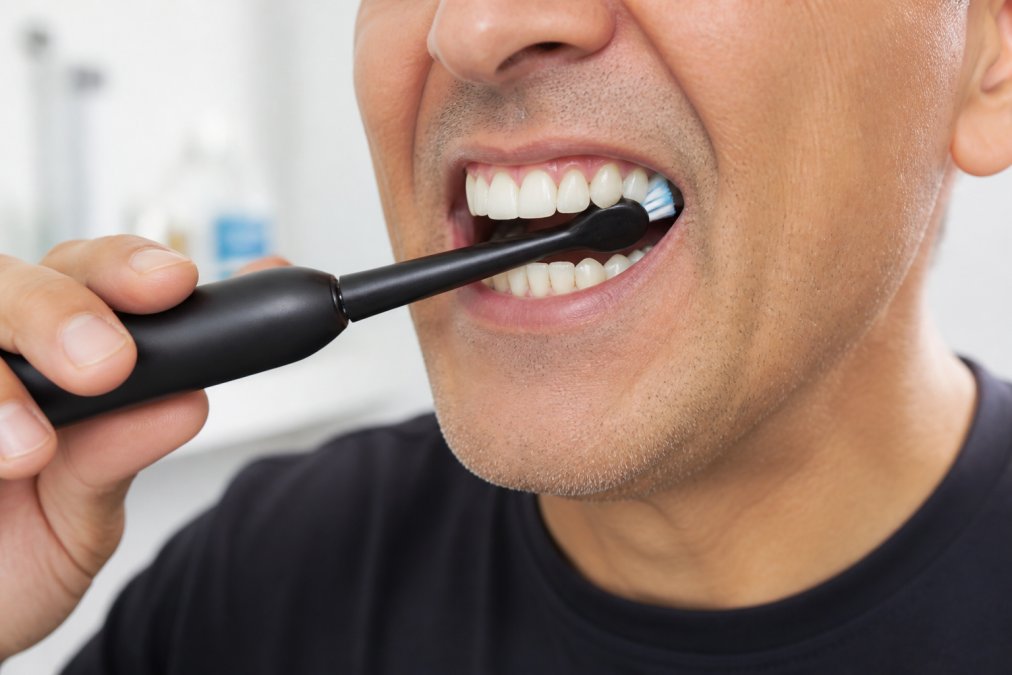Close-up of a person using an electric toothbrush with proper technique along the gum line, showing careful cleaning around implant area with soft bristles