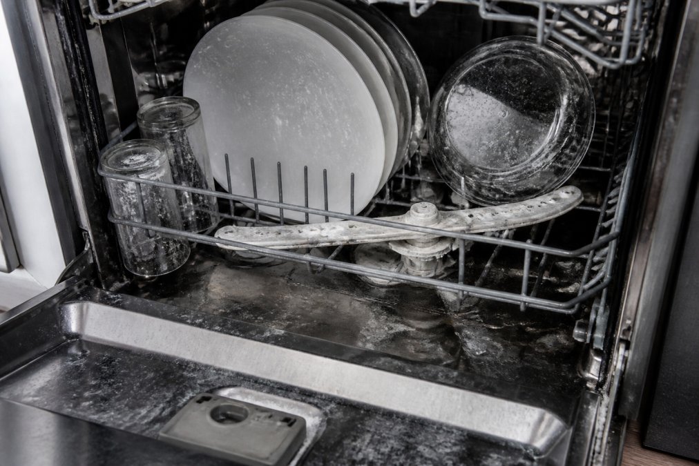 Close-up of a dishwasher interior showing white mineral spots and residue on clean dishes, with the spray arm visible and slightly corroded, door open to reveal hard water damage