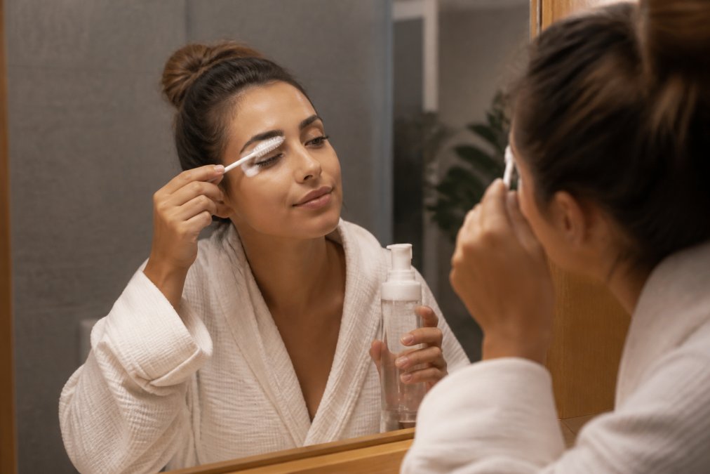 A woman gently cleaning her lash extensions with a soft brush and foam cleanser in front of a bathroom mirror, warm bathroom lighting illuminating her face as she demonstrates proper lash bath technique