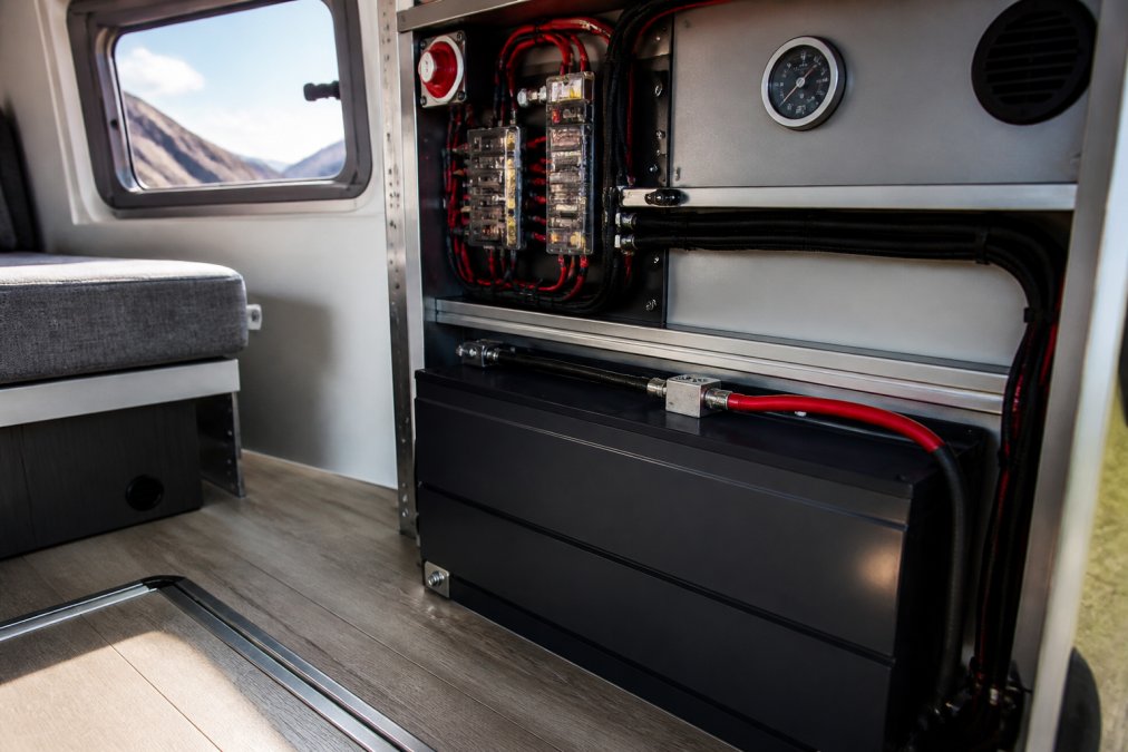 Close-up of a lithium battery mounted in a campervan installation with temperature gauge and battery management system display visible on the nearby control panel, natural daylight streaming through a small window
