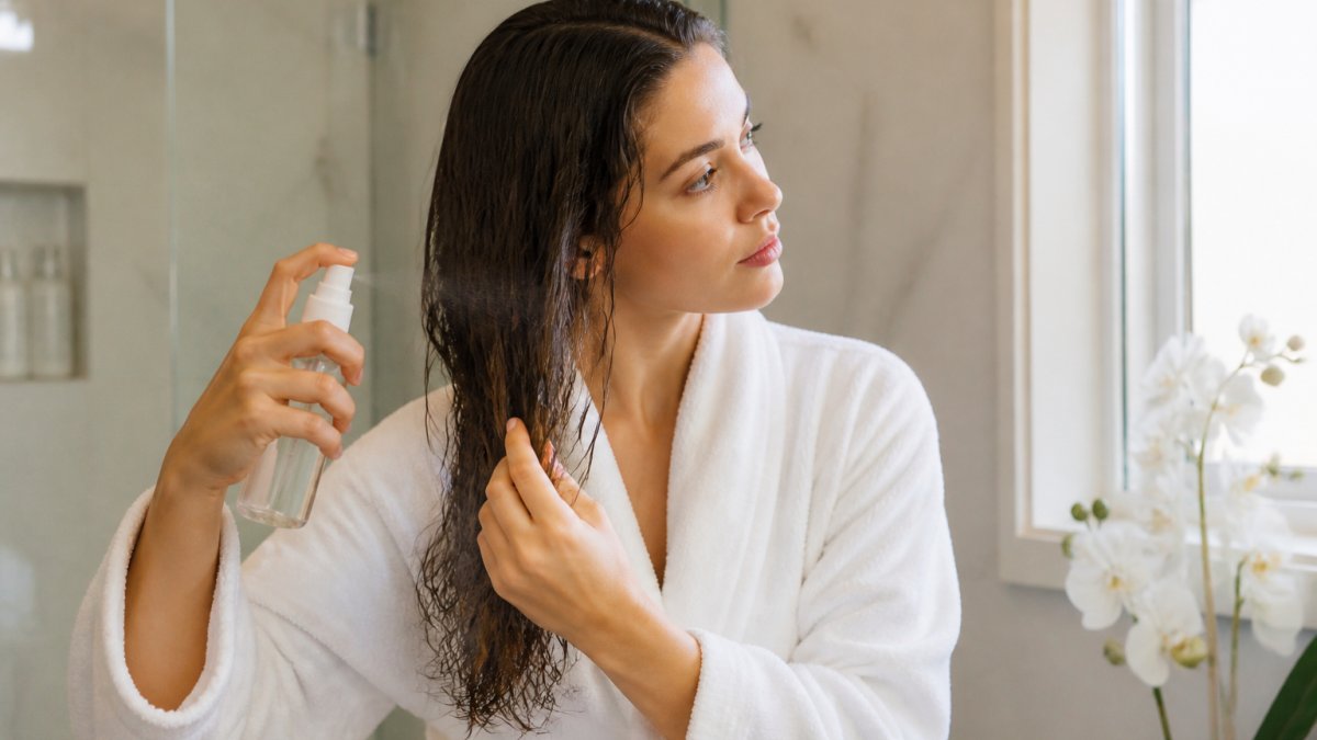 Woman applying heat protectant spray to damp hair sections, demonstrating even distribution from roots to ends in natural bathroom lighting