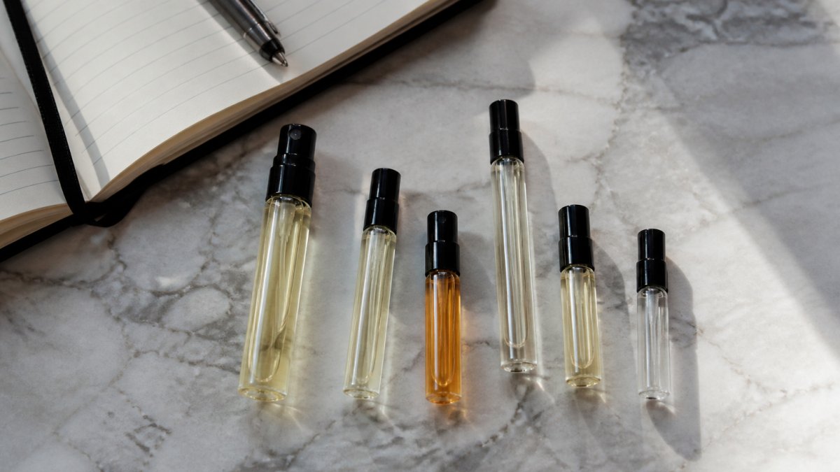 Small glass perfume sample vials arranged on a marble surface next to a notebook and pen in soft natural light