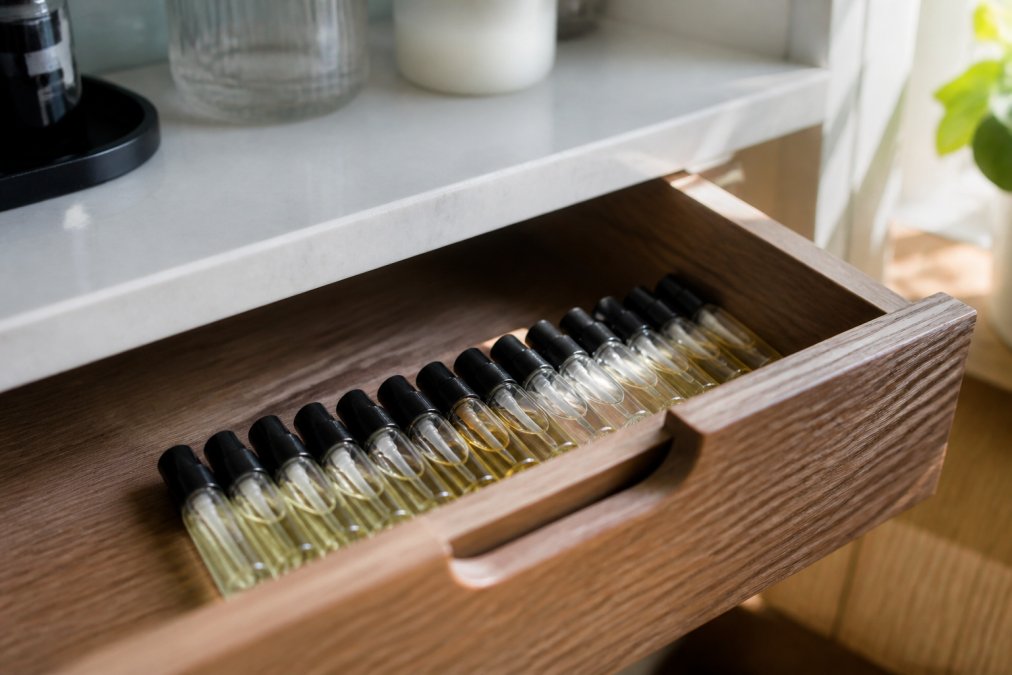 A neat row of small glass perfume vials arranged in a cool, shaded drawer away from a sunlit window, soft natural light falling on the wooden drawer interior