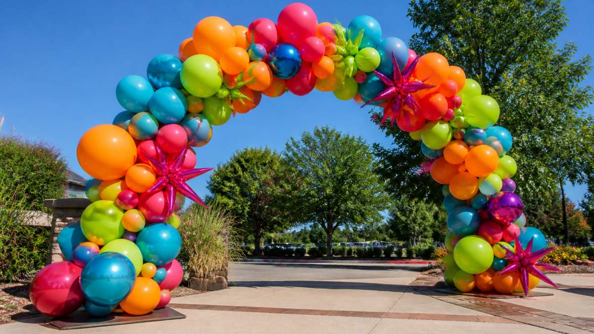 Professional balloon arch installed outdoors in bright sunlight with vibrant latex and foil balloons against clear blue sky and green trees in background