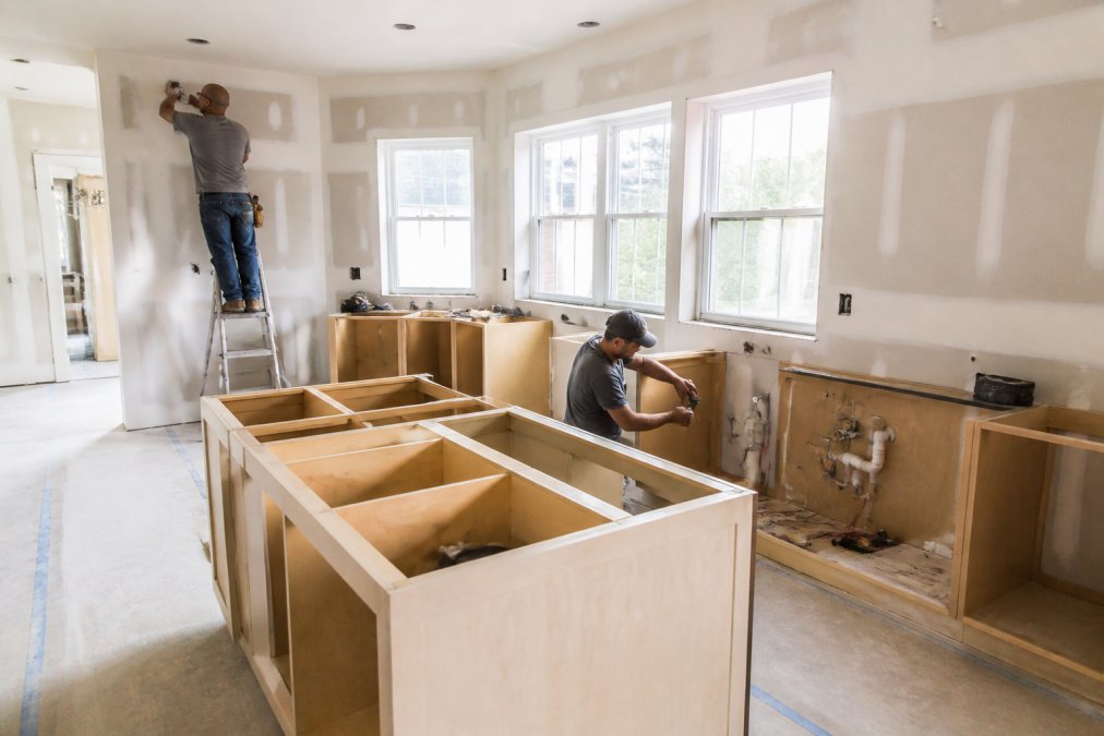 Interior of a kitchen under active renovation showing exposed drywall, open cabinetry, and contractors working on plumbing and electrical installations with natural light streaming through windows