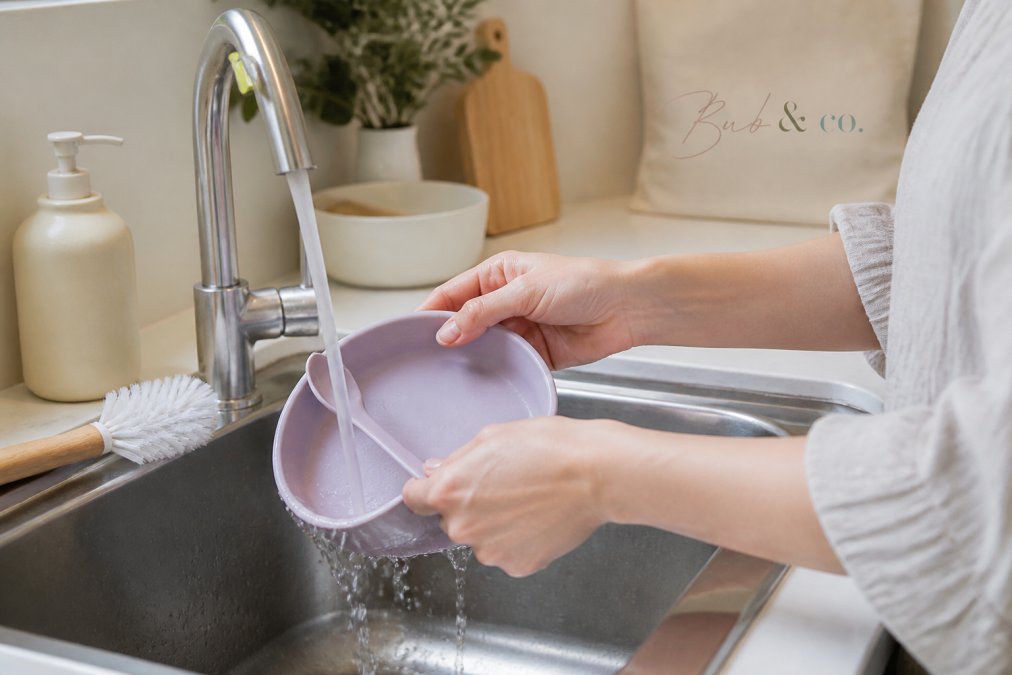 Parent washing a pastel silicone baby plate and spoon with mild soap in a bright kitchen sink