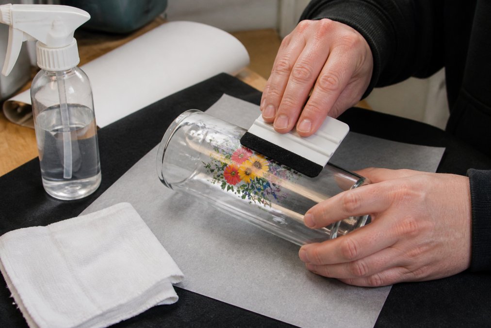 Close-up of hands using a squeegee to press a UV DTF decal onto a glass tumbler on a clean workbench, with isopropyl alcohol spray bottle and lint-free cloth nearby