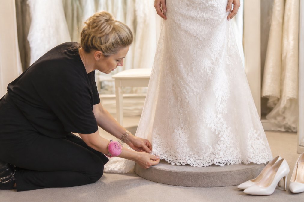 A seamstress carefully pinning the hemline of a white wedding gown on a bride standing on a fitting platform in a bright bridal boutique, with wedding shoes and a satin sash visible nearby