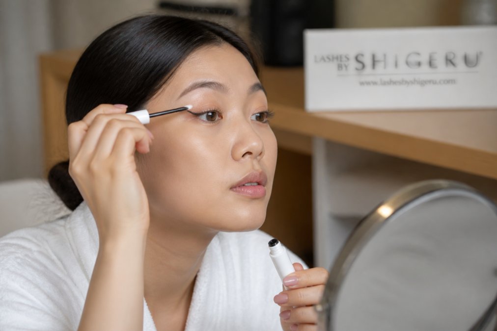 Person carefully applying a lash serum or growth product to their lash line using a small brush applicator, demonstrating proper aftercare technique with steady hands and good lighting