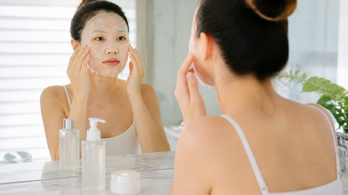 A young woman in a bright bathroom pressing a sheet mask onto her face, with toner and moisturizer bottles on the marble counter, soft morning light