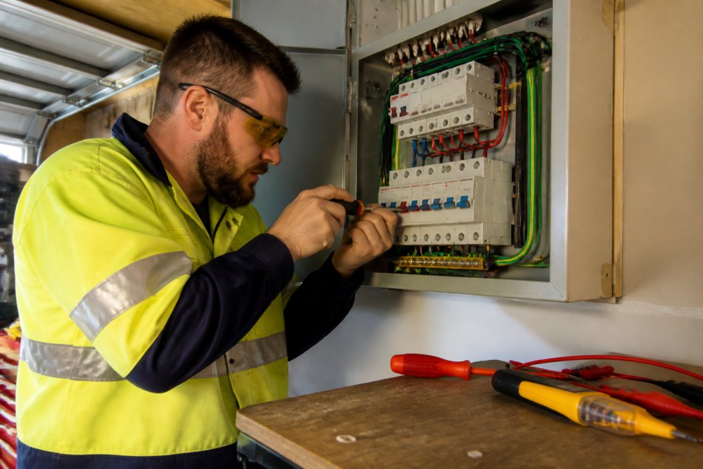 A qualified electrician in high-visibility work gear carefully installing circuit breakers inside an open residential switchboard, with screwdrivers and a voltage tester laid out on a nearby workbench, under bright garage lighting